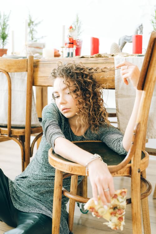 Free A woman with curly hair sits alone indoors, holding a slice of pizza, embodying holiday loneliness. Stock Photo
