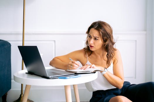 A woman in a white top attentively takes notes using a laptop at home.