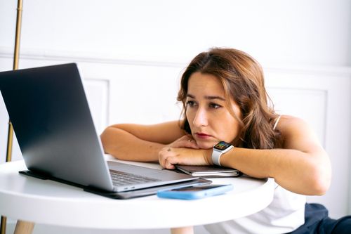 Free Woman appears tired while looking at a laptop, leaning on a table, in a minimalist indoor setting. Stock Photo