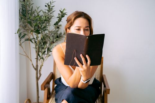 Free A woman in white top and smartwatch enjoys reading indoors on an armchair beside a plant. Stock Photo