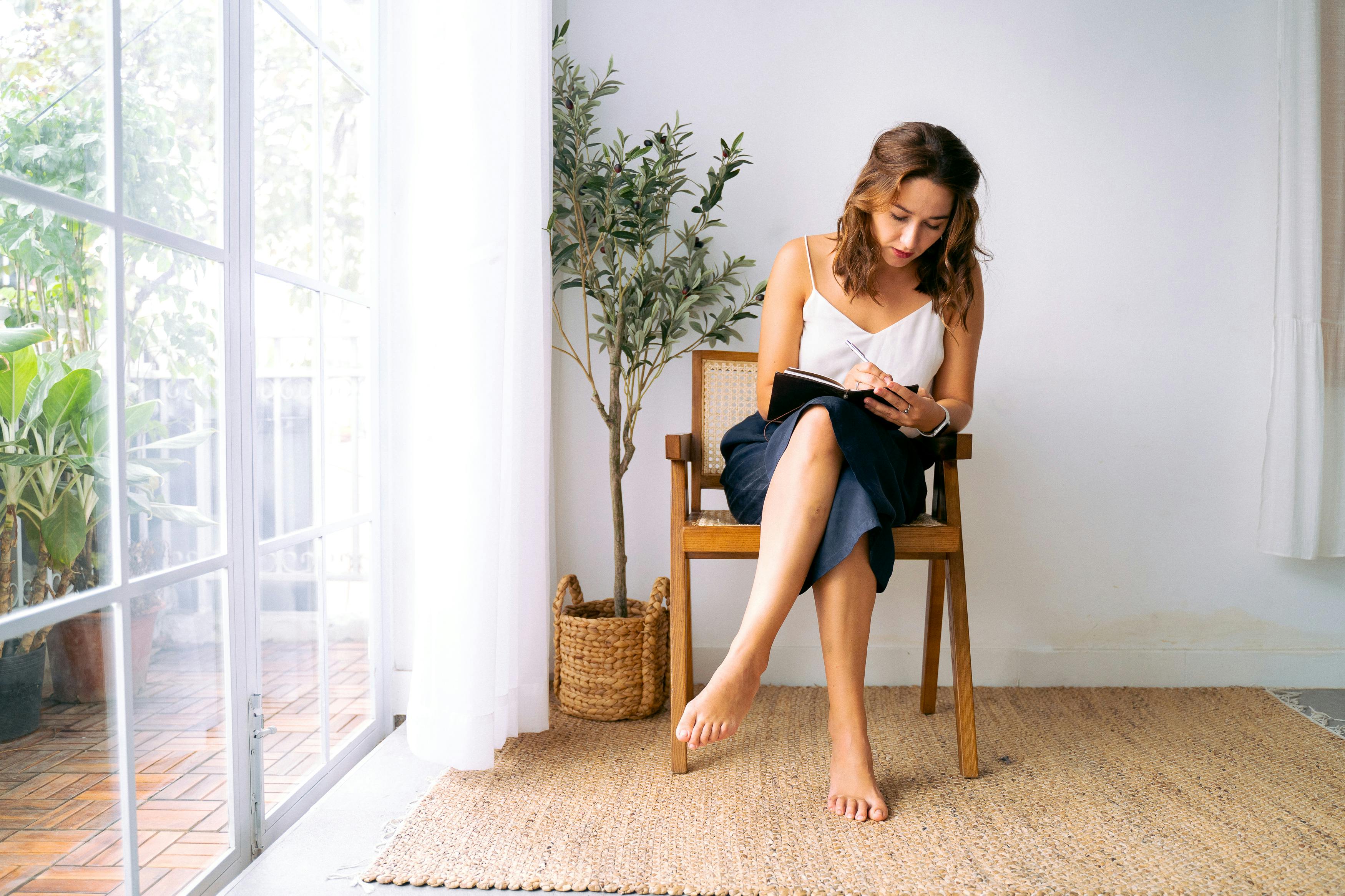 A woman sits barefoot in a cozy chair, taking notes in a sunlit room with potted plants.