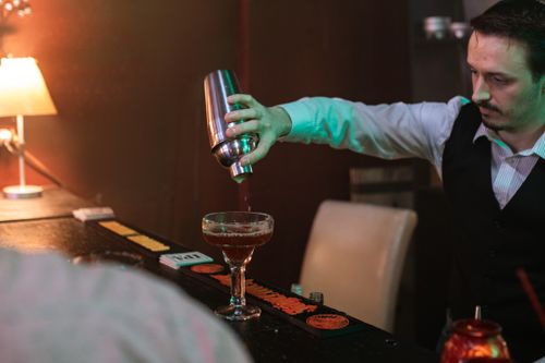 Free A bartender skillfully pours a cocktail from a shaker into a glass on a dimly lit bar counter. Stock Photo