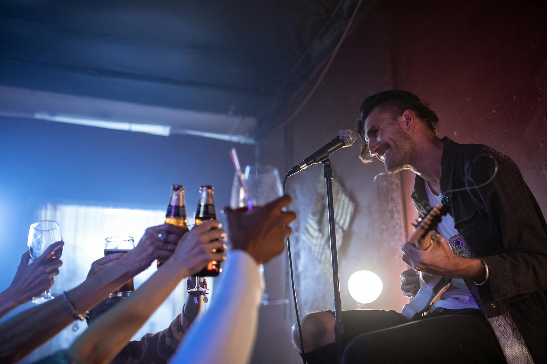 Musician playing guitar and singing on stage while audience cheers with drinks indoors.