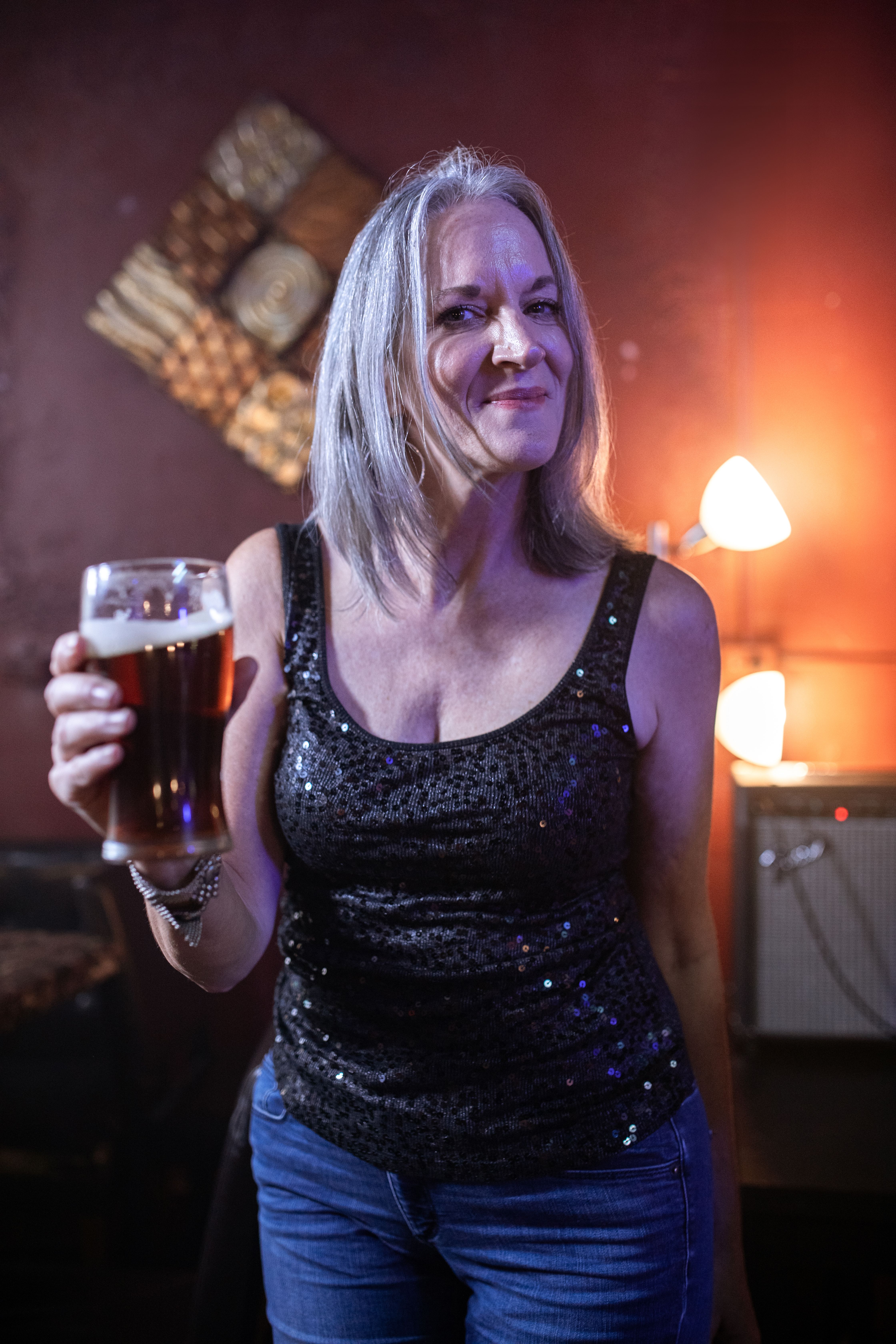 Elegant woman in a sequin top holding a glass of beer at a vibrant bar setting.