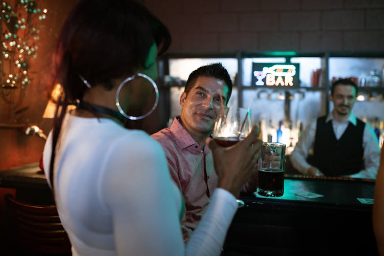 A Man Sitting At The Bar Counter Looking At A Woman Holding A Cocktail