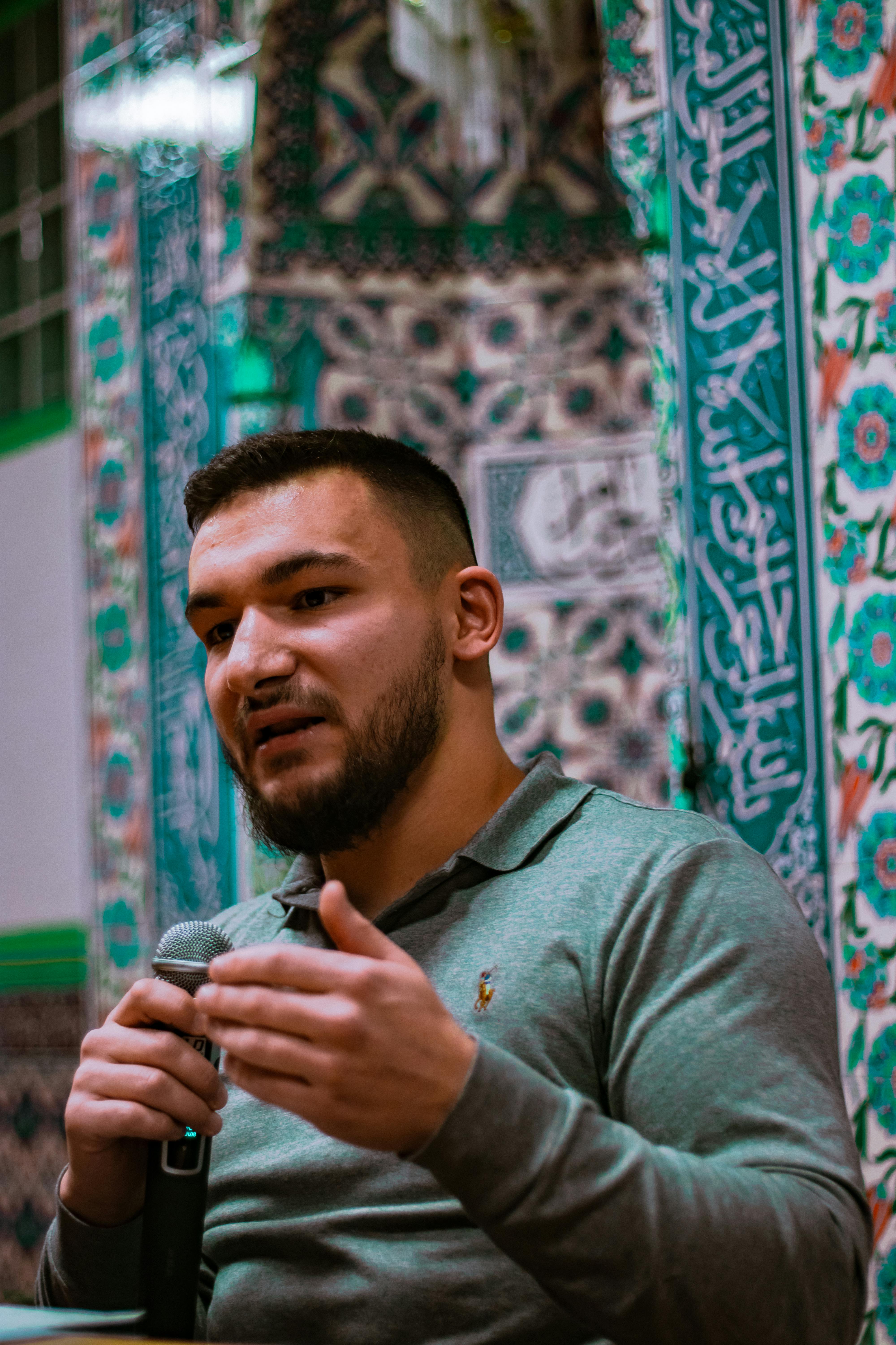 Bearded man holding a microphone, speaking in a decorative room with intricate patterned walls.
