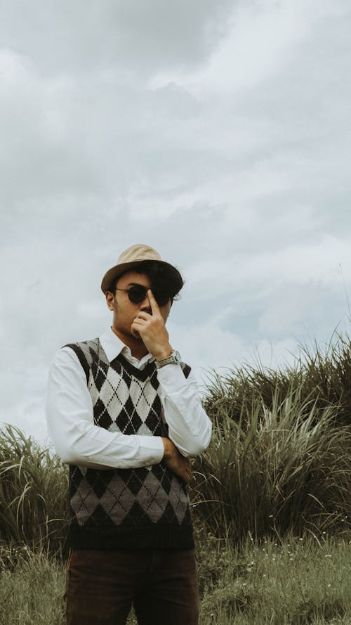 Serious young ethnic male in trendy wear and hat touching sunglasses while standing on grassy meadow in countryside on overcast day