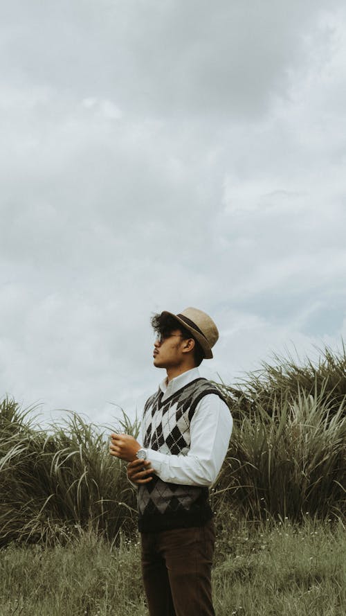 Side view contemplative young ethnic male in trendy clothes and hat standing on lush grassy field and looking away under cloudy sky