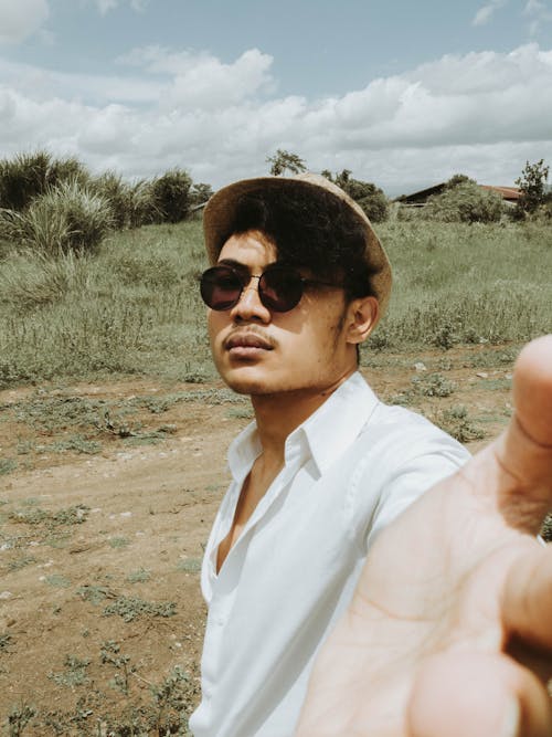 Emotionless young ethnic male in white shirt hat and trendy sunglasses touching camera while standing on grassy vast rural area