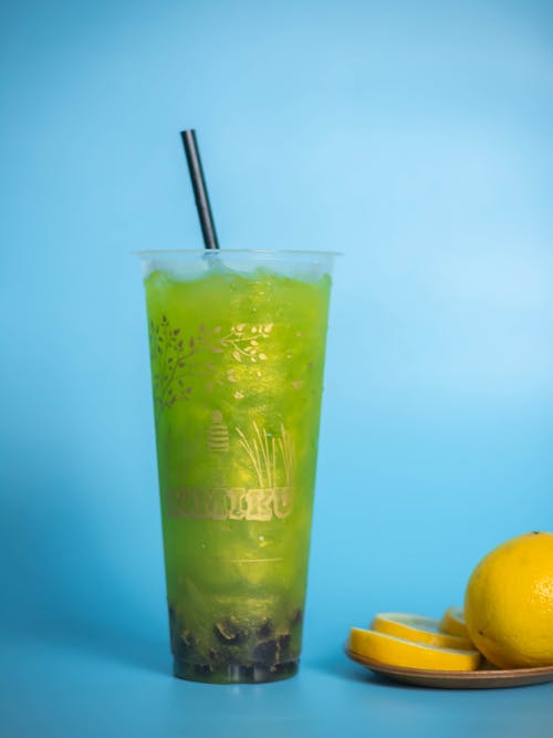 Plastic glass of cold yummy sour lemonade of green color with straw placed on blue background near saucer with lemon sliced