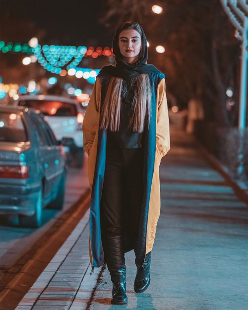 Free Young woman in vibrant coat walking on Mashhad sidewalk at night. Stock Photo