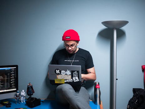 Freelancer in a red beanie working on a laptop indoors with decorative action figures and tech setup.