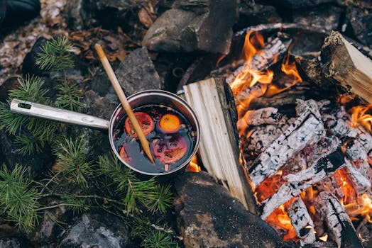 Pot of mulled wine simmering over a campfire outdoors with oranges and spices.