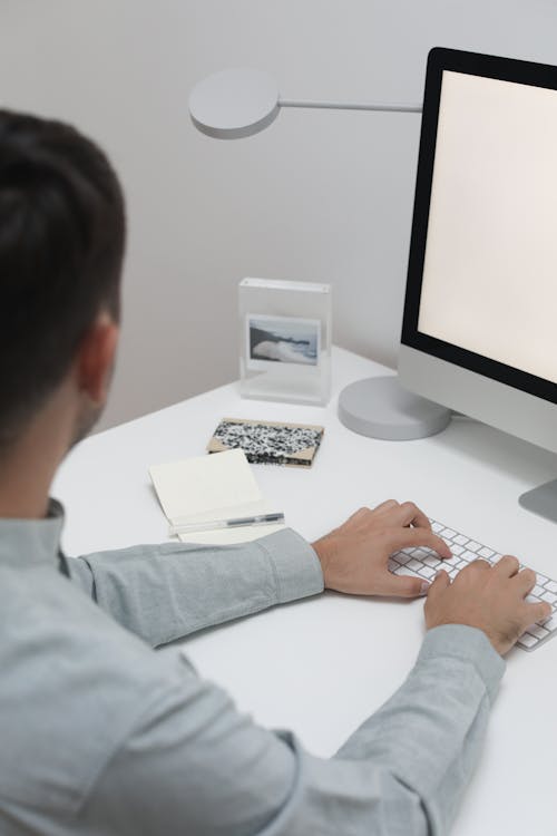 Crop anonymous male in formal outfit typing on keyboard while sitting at table with computer and notepads