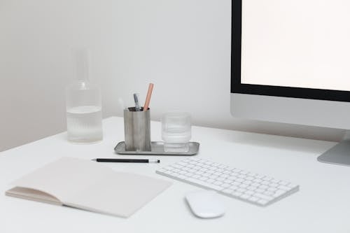 Modern computer with wireless keyboard and mouse on table with open notepad and bottle of water