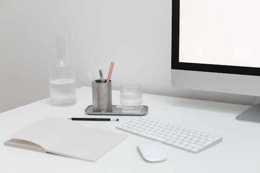 Modern computer with wireless keyboard and mouse on table with open notepad and bottle of water