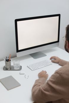 Crop anonymous male in sweater and eyeglasses sitting at table with computer and earbuds while working online