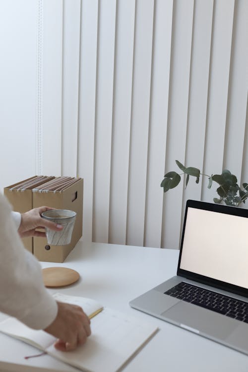 Crop faceless person standing near table with laptop and folders while drinking coffee