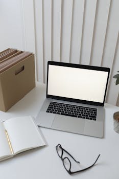 A minimalist office desk setup featuring a laptop, eyeglasses, a notebook, and a file box.
