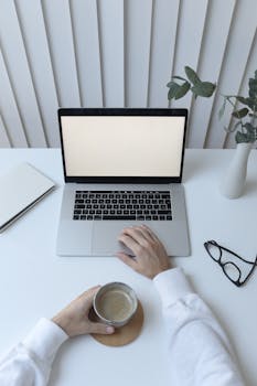 High angle of crop anonymous person browsing netbook while drinking coffee in modern workplace