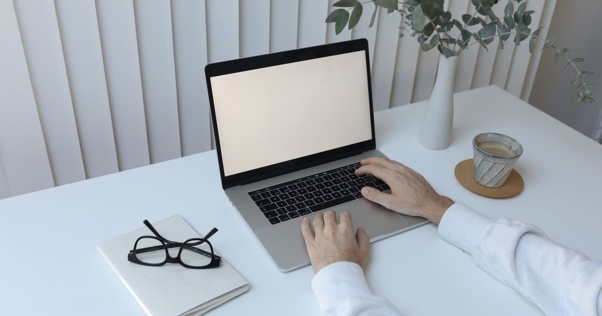 A person types an email reply on a laptop at a tidy white desk with coffee, glasses, and a plant
