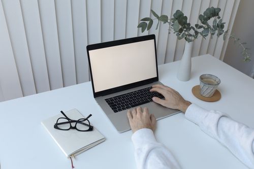 Free High angle of crop faceless person sitting at table with cup of hot coffee and notepad while working on netbook Stock Photo
