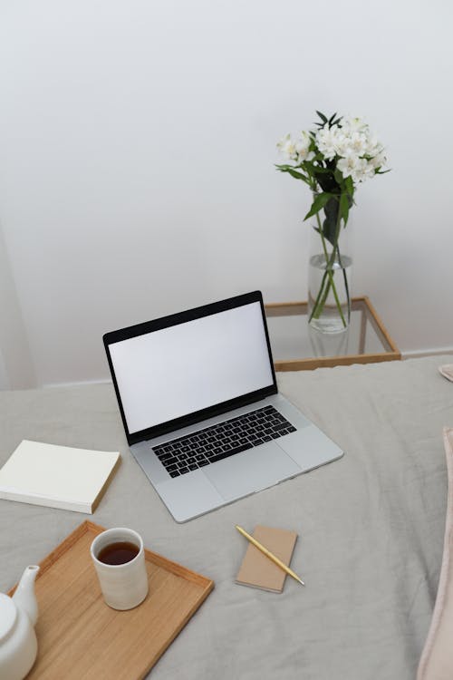 From above of opened laptop with empty screen and book placed on comfortable bed with coffee set and notebook near small table with flowers in vase
