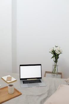A clean, minimalist home workspace featuring a laptop, coffee, and flowers on a bed.