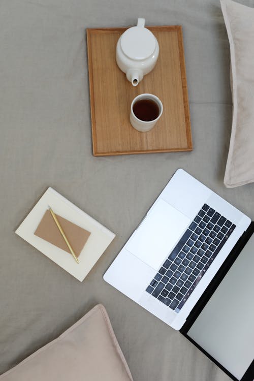 Top view of opened laptop with white screen and wooden tray with coffee set placed on comfortable bed with notebook and pen during distance lesson at home