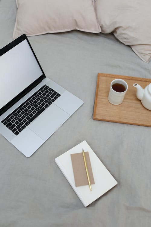 Free From above of book with jotter and pen arranged on comfortable bed with opened laptop and tray with coffee cup and pot during freelance work Stock Photo