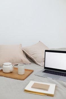 Wooden tray with tea cup and pot placed on bed with modern laptop and notebooks during remote work at home