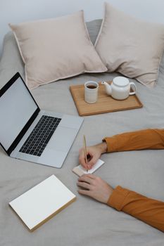 From above of crop unrecognizable male student taking notes in jotter while studying on bed with book and laptop during distance lesson