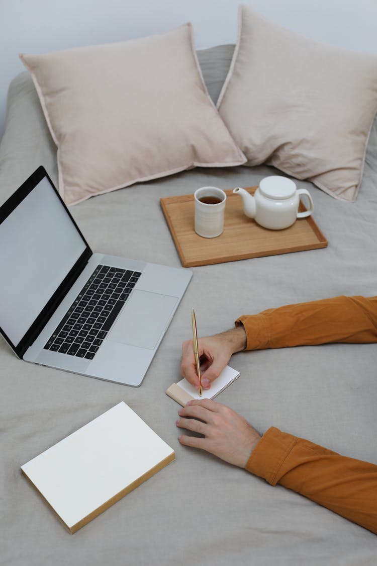 Faceless Guy Writing In Notebook On Bed With Laptop During Online Studies