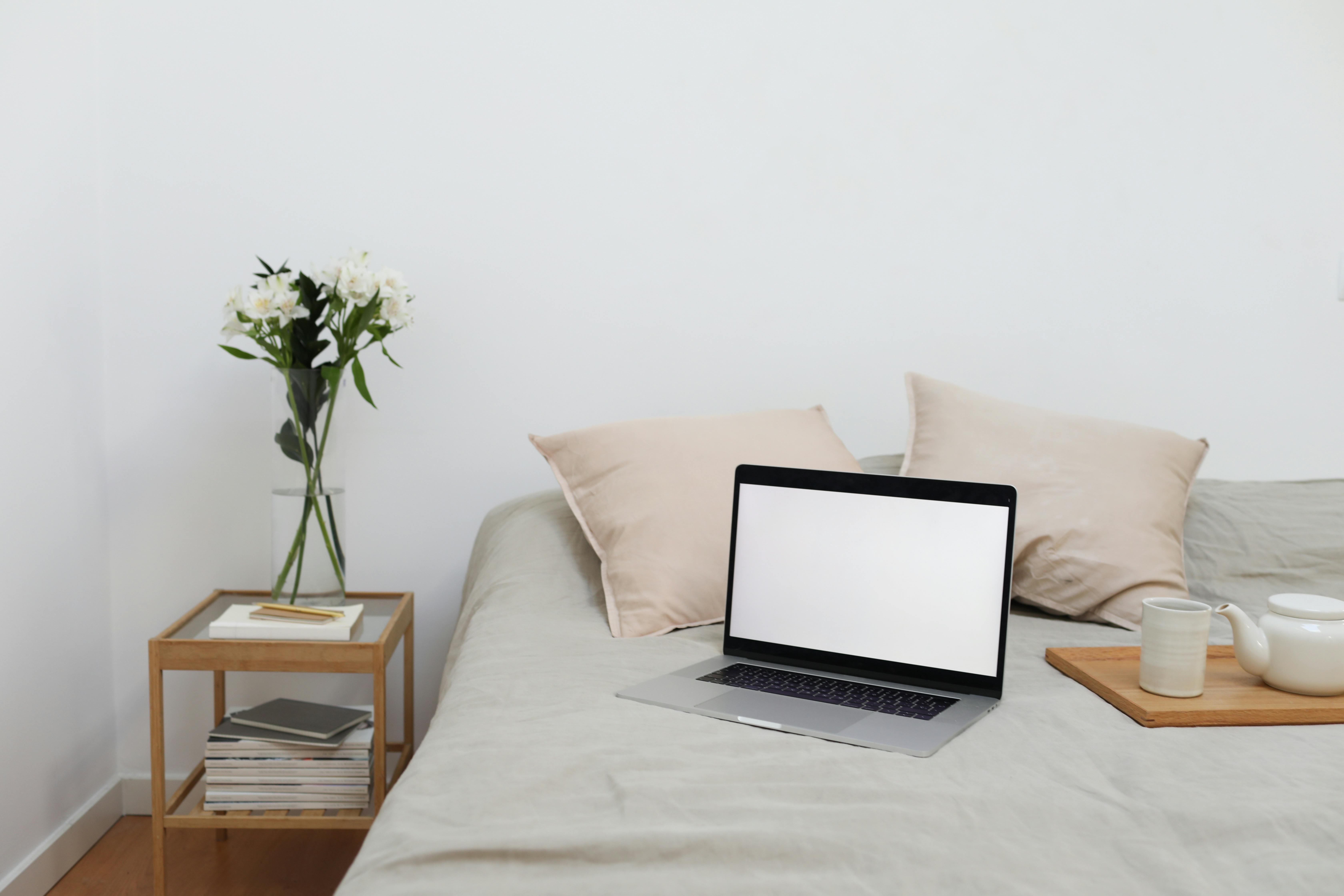 tray with tea set and laptop placed on bed near table with flowers vase