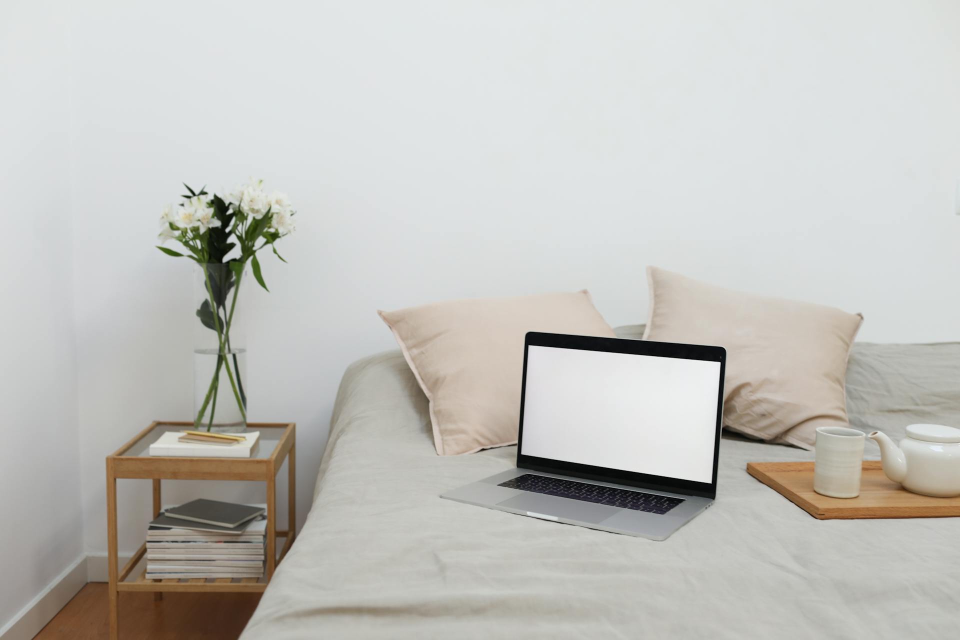 Tray with tea set and laptop placed on bed near table with flowers vase