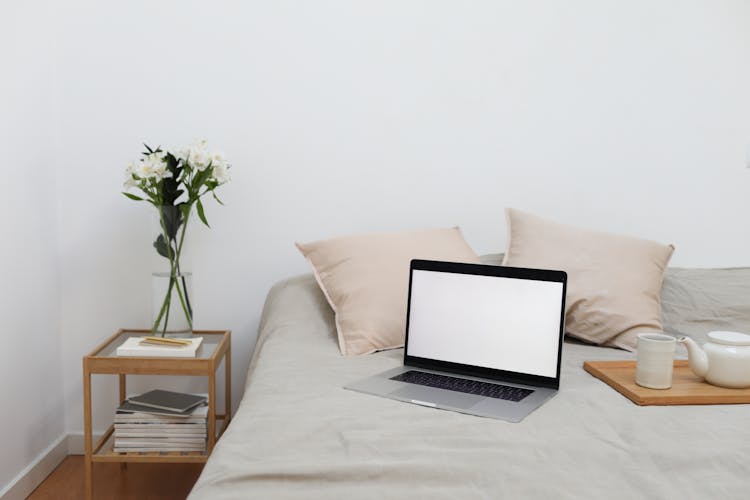 Tray With Tea Set And Laptop Placed On Bed Near Table With Flowers Vase