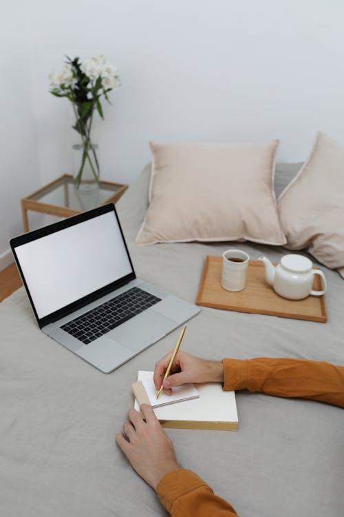 Free From above of crop faceless young male freelancer taking notes in planner while working remotely on laptop on bed with cup of aromatic coffee Stock Photo