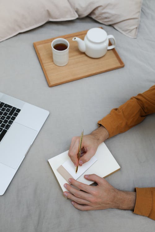 Free From above of crop anonymous self employed man taking notes in planner while working distantly on netbook on comfortable bed with cup of coffee Stock Photo