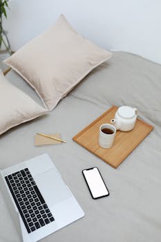 A comfortable home setup featuring a laptop, smartphone, and tea set on a bed, ideal for remote working.