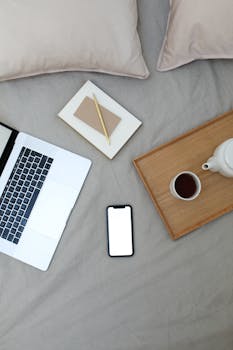 Top view of mobile phone with empty screen and planner placed on bed near opened netbook and wooden tray with coffee cup and pot during online work