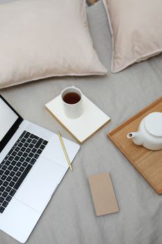 High angle of coffee cup and pot arranged on comfortable bed near opened laptop during remote work
