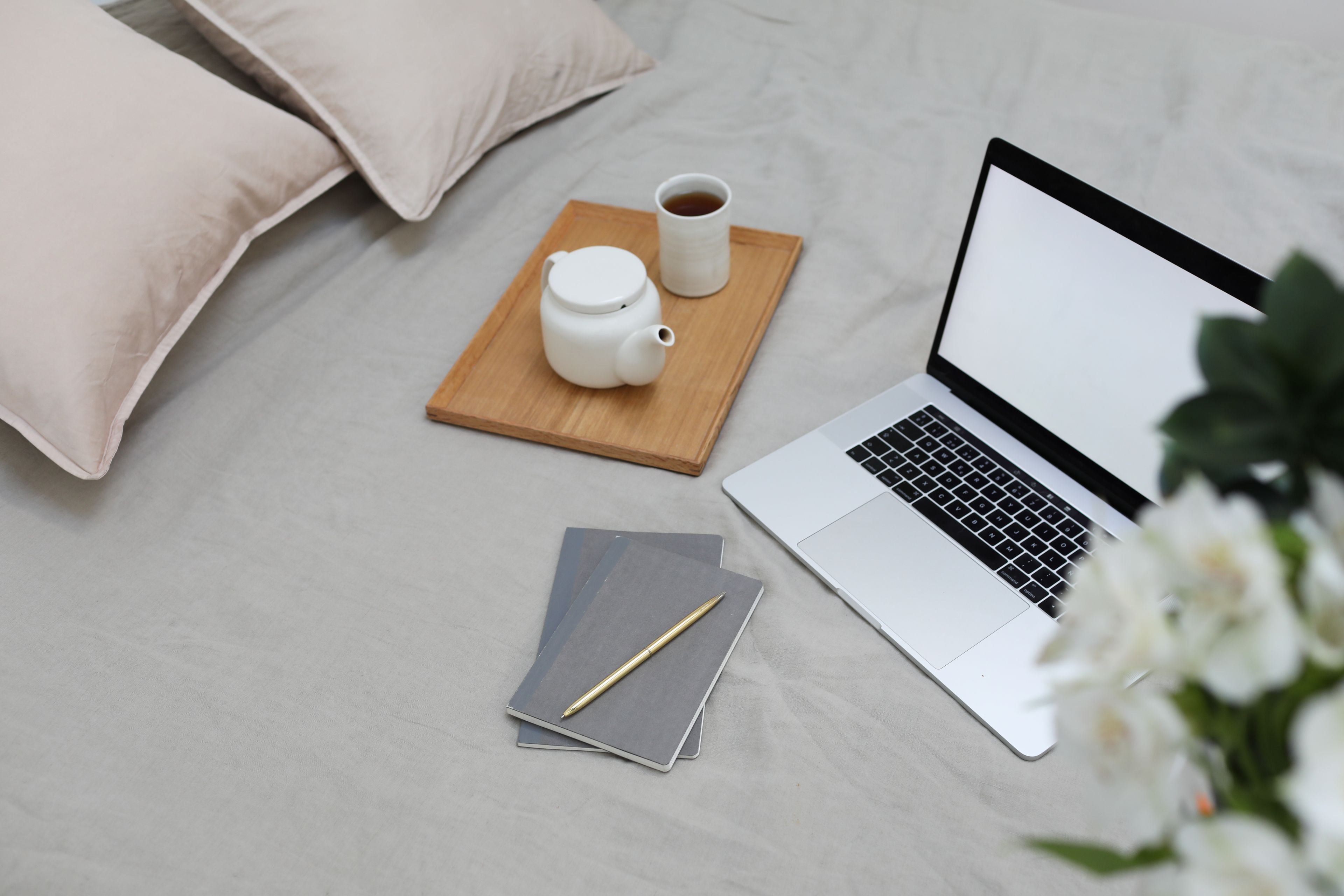 From above of wooden tray with coffee cup and pot placed on soft bed with notebooks and opened laptop with blank screen during distance studies
