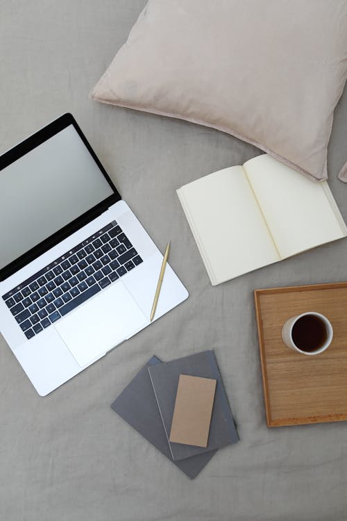 Free Top view of opened book and modern laptop with blank screen placed on bed near wooden tray with coffee cup during freelance work at home Stock Photo