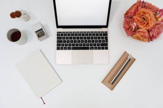 A top view of a minimalistic workspace featuring a laptop, flowers, and stationery on a clean desk.
