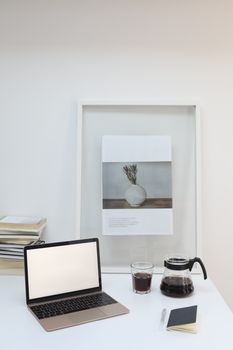 Opened laptop with empty screen placed near notebook and glass of fresh coffee on white table in light room