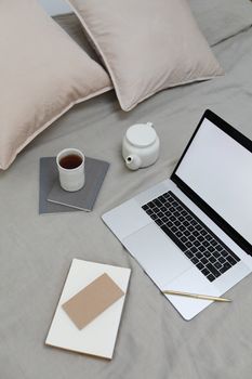 From above of laptop with blank screen placed near notebook and cup of hot tea on comfortable bed in bedroom in daytime
