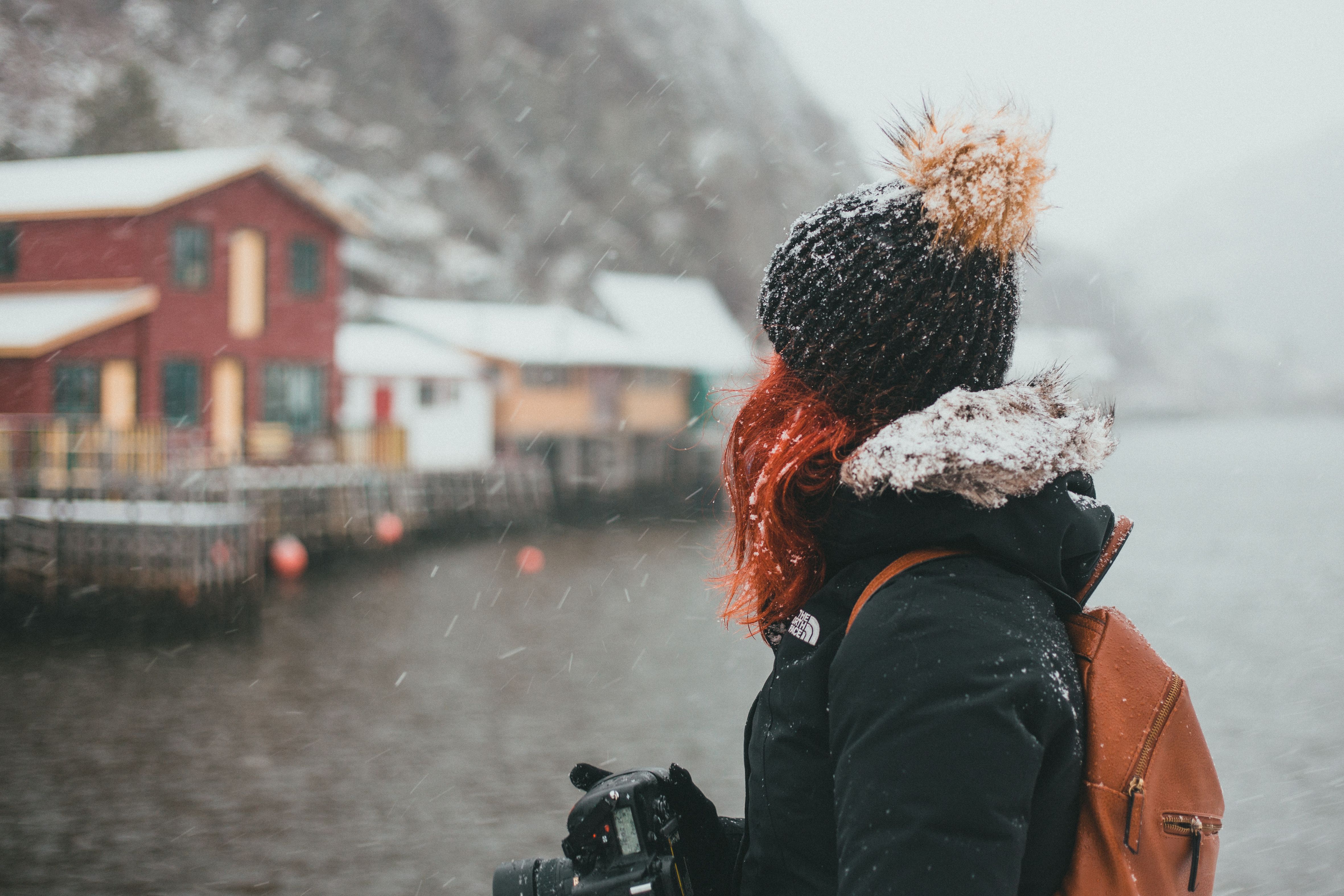 A traveler in winter clothes gazes at snowy cabins by a lake, capturing the serene winter landscape.