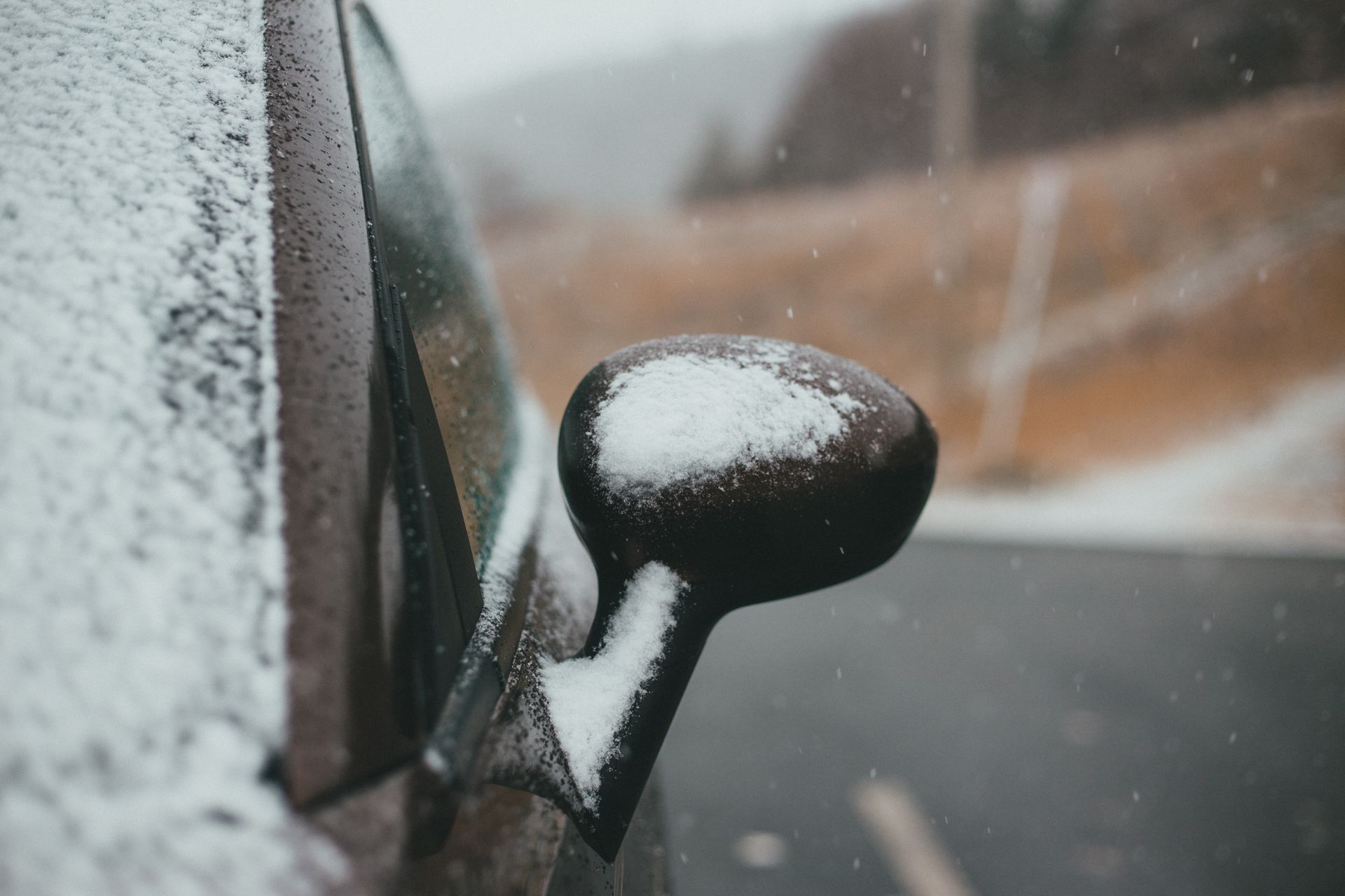 Close-up of a snow-covered car mirror on a rural road in winter, emphasizing cold weather travel.