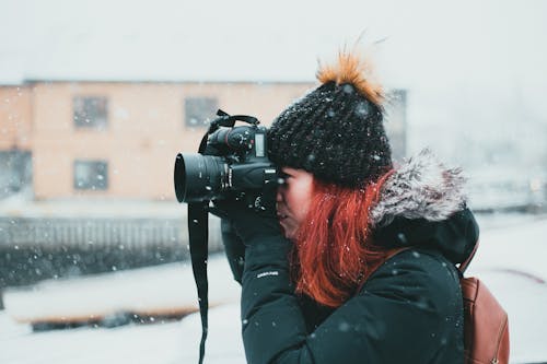 Side view of female photographer wearing warm outerwear taking pictures on professional photo camera during snowfall