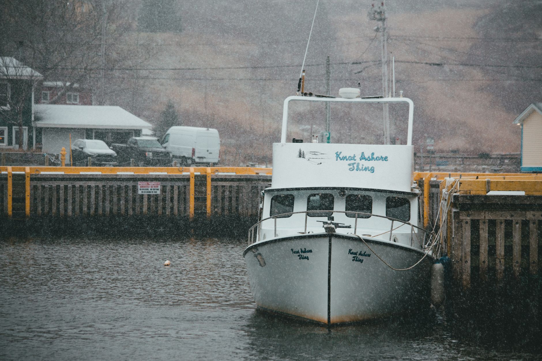 A boat named 'Knot Ashore Thing' docked at a harbor during snowfall, depicting a serene winter scene.
